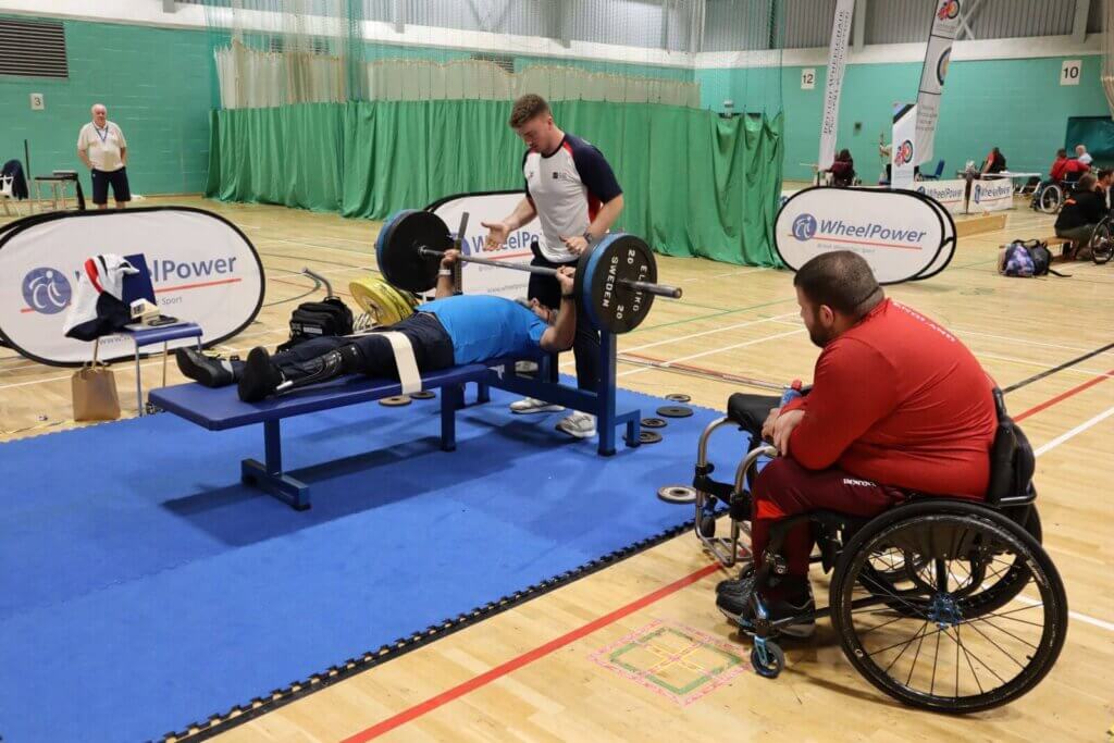 Mo listing weights on a benchpress with a spotter and trainer