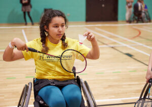 Young female wheelchair user playing badminton in an indoor badminton court.